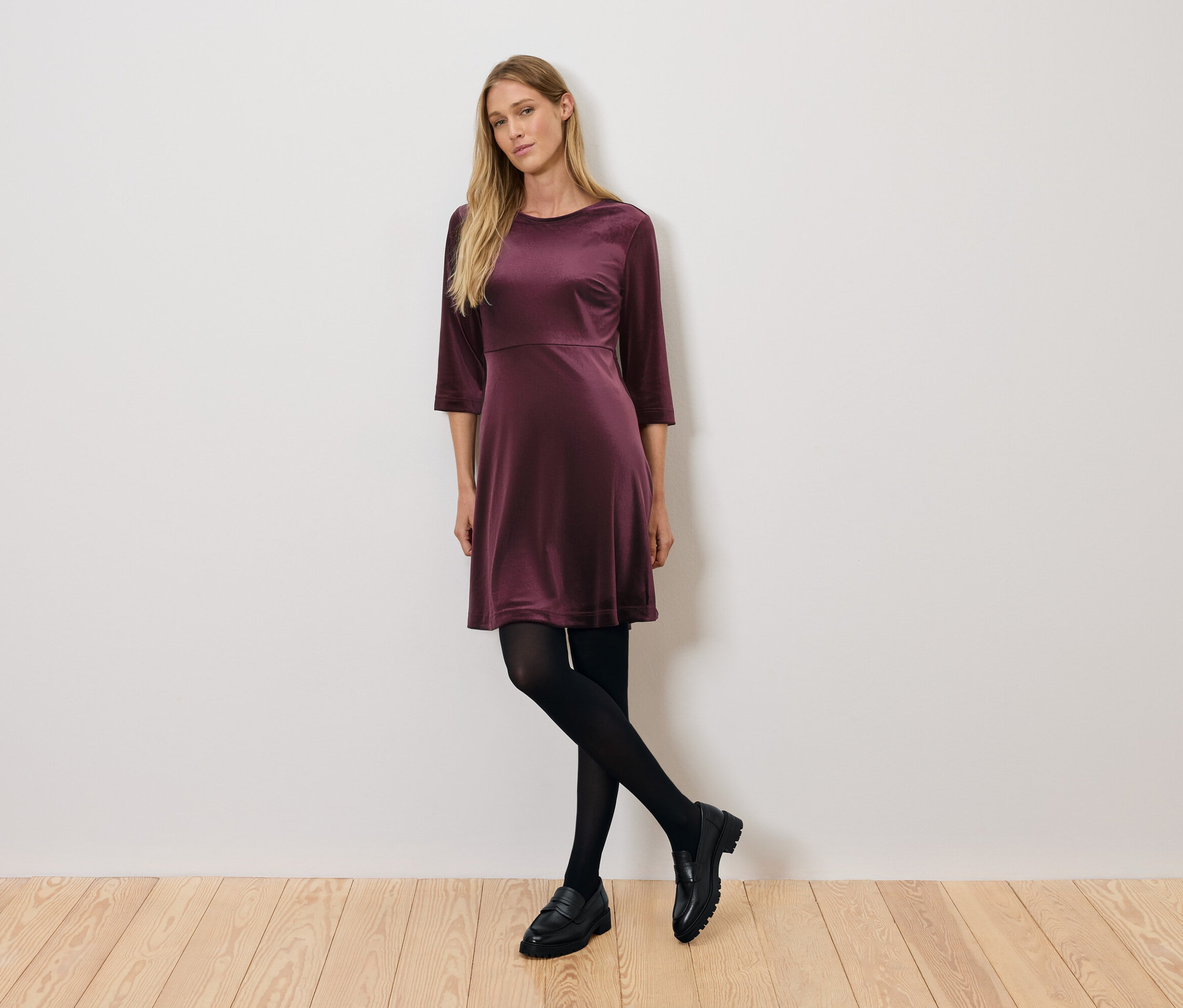 Une femme pose dans une robe de velours bordeaux à manches trois-quarts, des collants noirs et des chaussures noires.