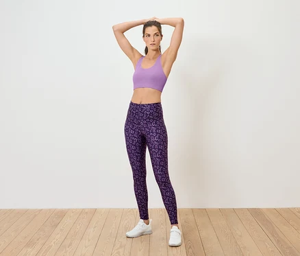 Une femme pose sur le sol en soutien-gorge de sport lilas et leggings violets.