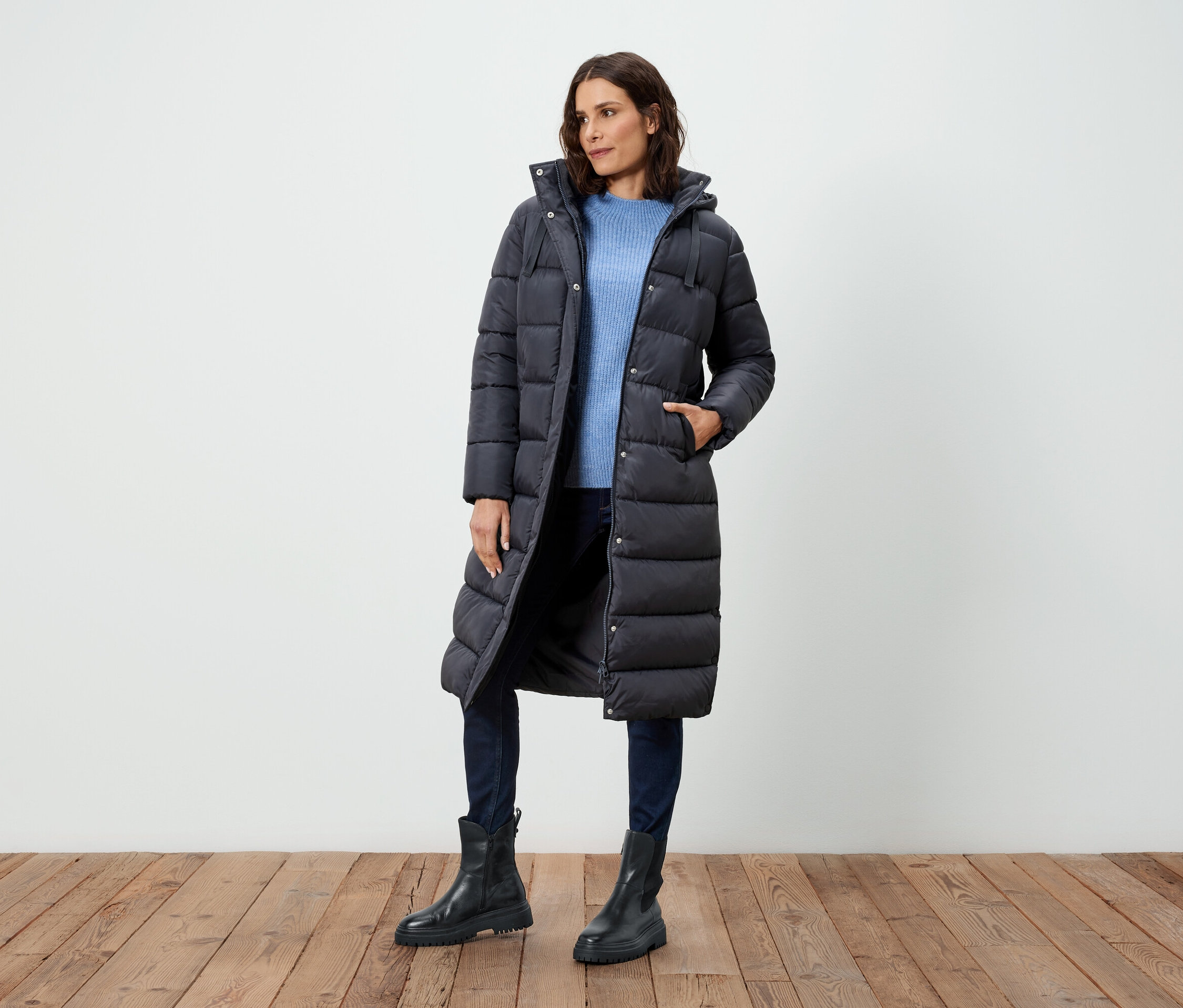 Femme posant dans un long manteau en duvet noir, un pull bleu, un jean et des bottes noires.