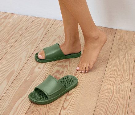 Une personne se tient pieds nus sur un plancher en bois à côté de deux paires de tongs vertes.