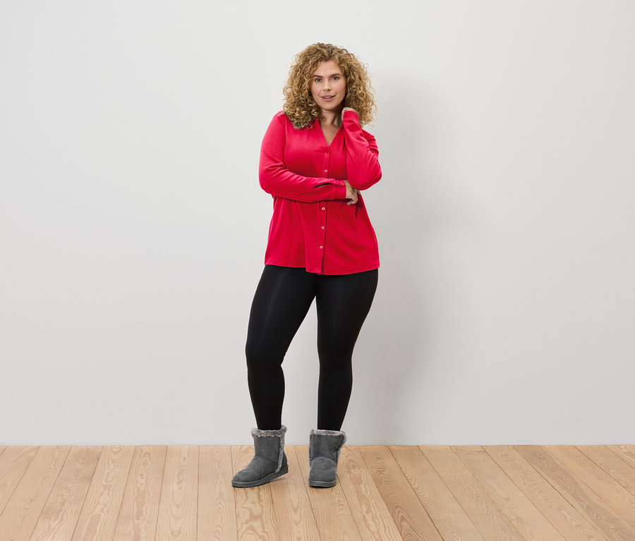 Une femme aux cheveux bouclés pose devant un mur blanc, portant un chemisier rouge, un legging noir et des bottes grises.