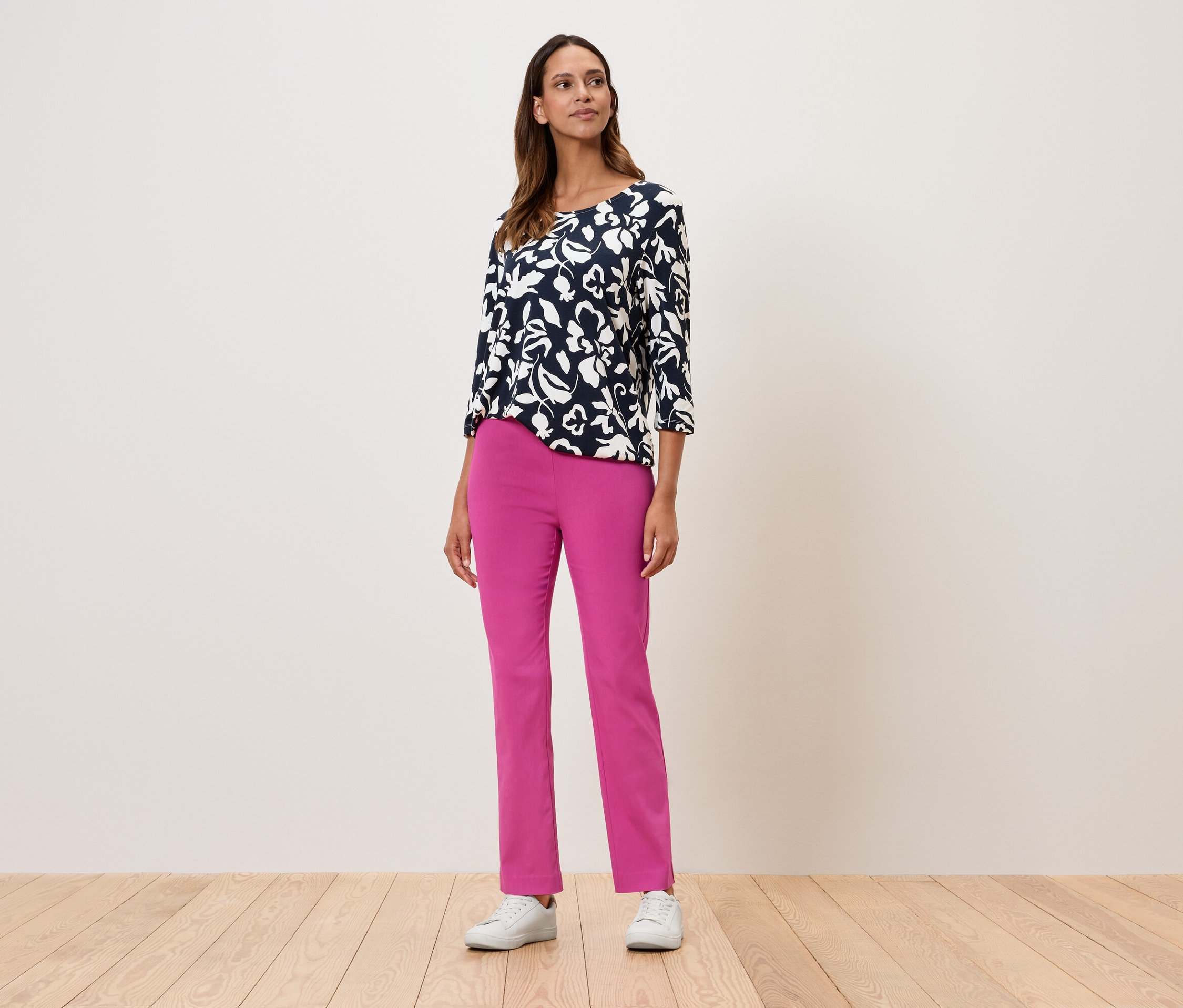 Une femme debout portant un haut à fleurs à manches trois-quarts, un pantalon rose et des baskets blanches.