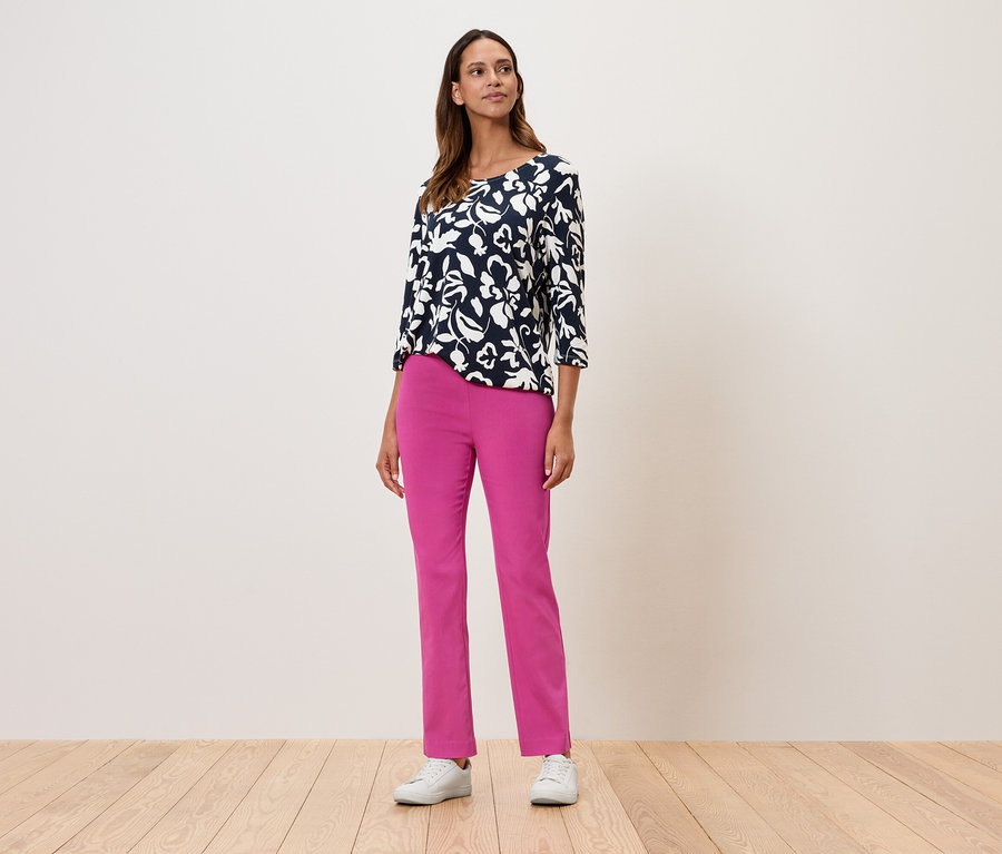 Une femme debout portant un haut à fleurs à manches trois-quarts, un pantalon rose et des baskets blanches.