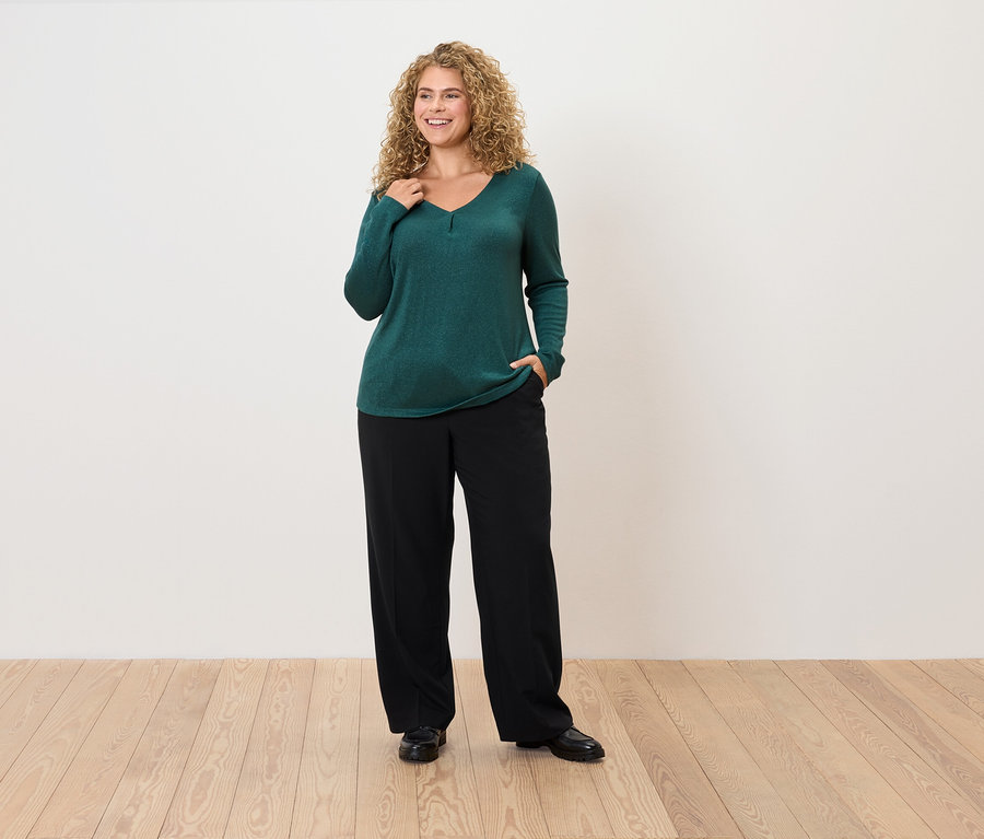 Une femme aux cheveux bouclés pose dans un pull vert émeraude et un pantalon noir.