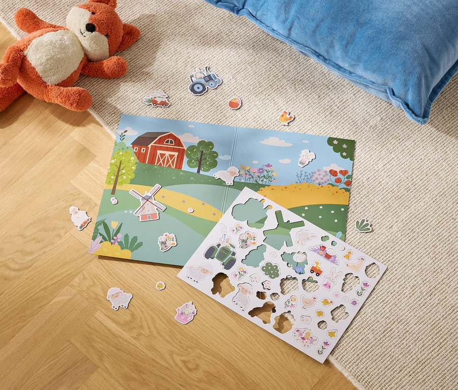 Un renard en peluche est allongé à côté d'un livre ouvert avec une illustration de ferme et une feuille d'autocollants de ferme sur un sol recouvert de moquette.