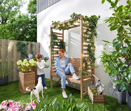 Une femme est assise sur un banc de jardin avec une arche de roses tandis qu'un enfant joue avec un ballon. Une guirlande lumineuse solaire « Ampoules » est suspendue au-dessus du banc. Dans le jardin, il y a 2 colonnes à plantes « Tinus » et un bac à herbes avec support « Elin ».