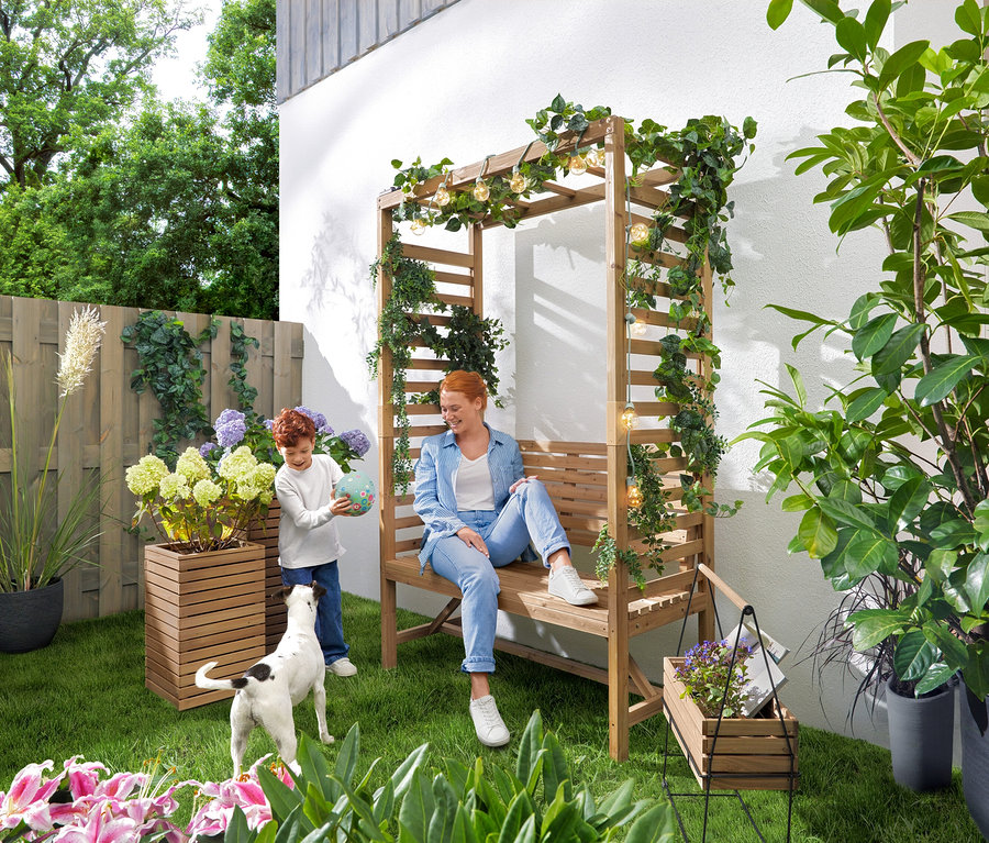 Une femme est assise sur un banc de jardin avec une arche de roses tandis qu'un enfant joue avec un ballon. Une guirlande lumineuse solaire « Ampoules » est suspendue au-dessus du banc. Dans le jardin, il y a 2 colonnes à plantes « Tinus » et un bac à herbes avec support « Elin ».