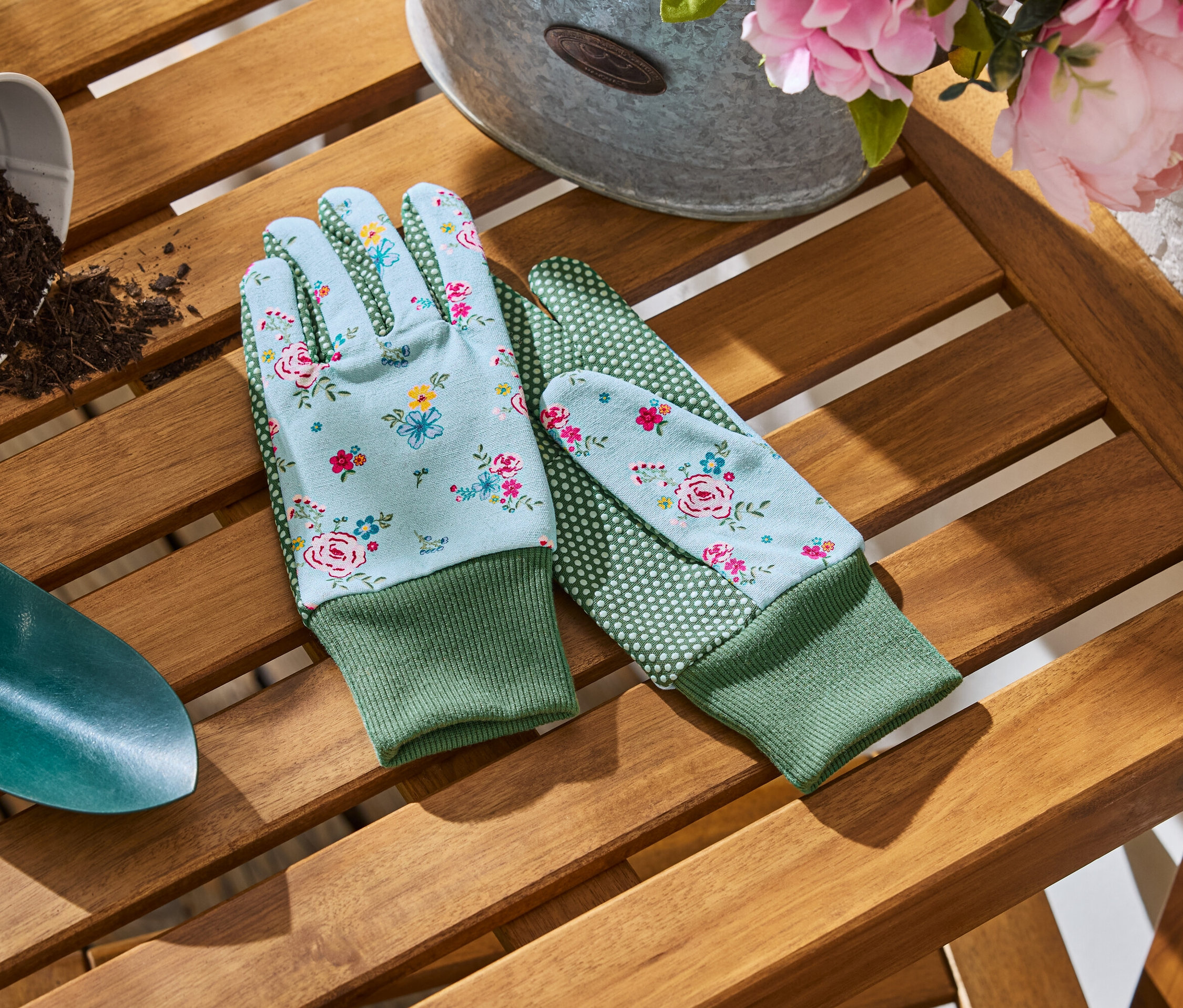 Sur une table en bois se trouvent des gants de jardin à motifs floraux, une petite pelle et un pot de terre.