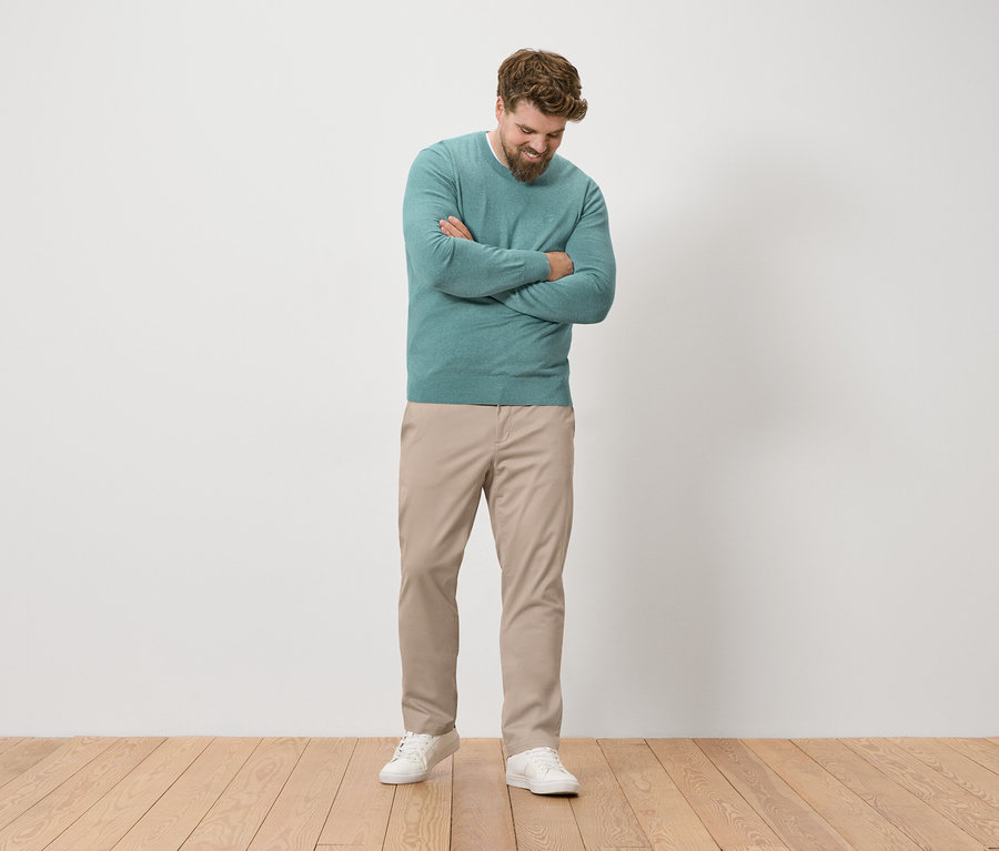 Un homme barbu se tient debout, les bras croisés, portant un pull vert, un pantalon beige et des baskets blanches.