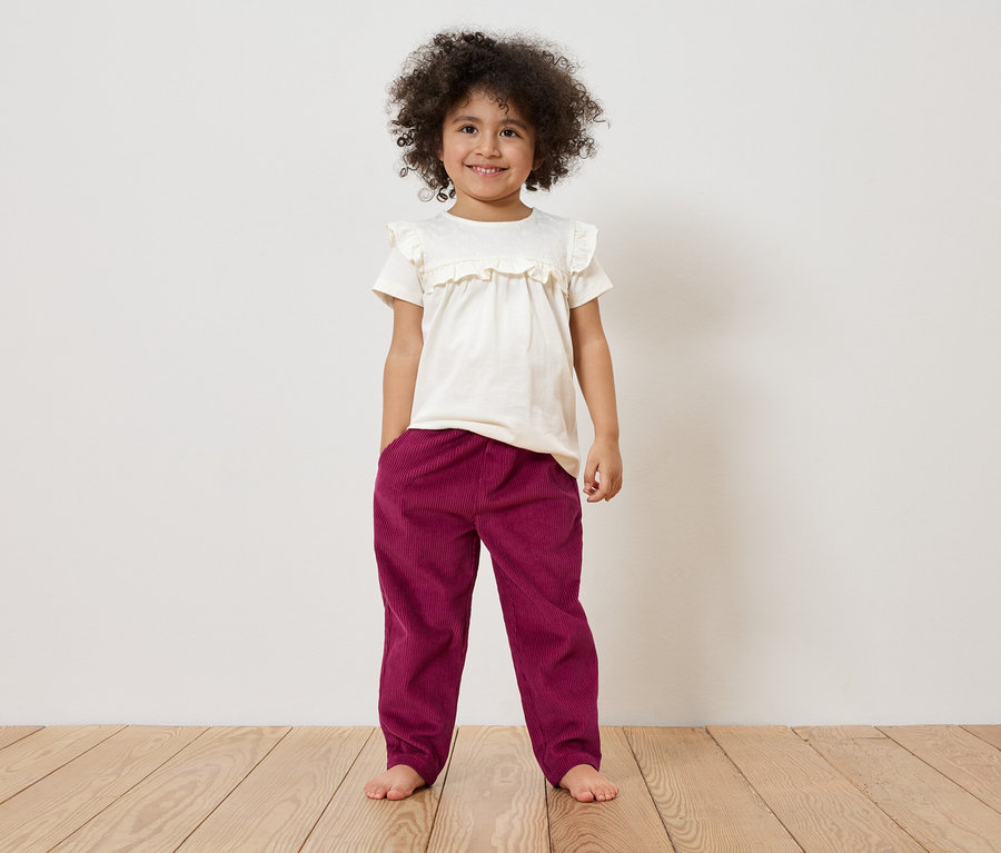 Une fillette aux cheveux bouclés se tient pieds nus devant un mur blanc, portant un t-shirt blanc et un pantalon bordeaux.