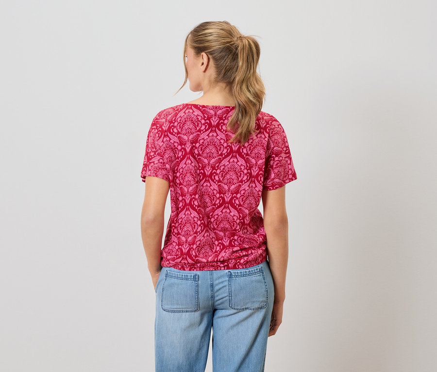 Une femme aux cheveux blonds porte un t-shirt rouge à motifs et un jean bleu.