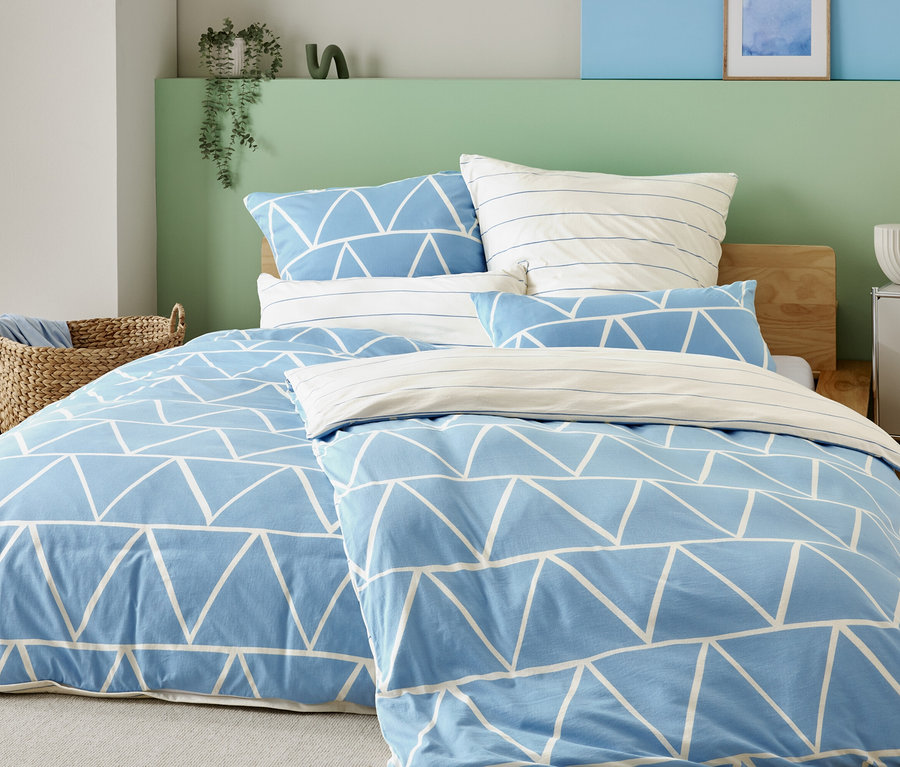 Lit avec housse bleu clair à triangles blancs et taies d'oreiller rayées, à côté d'un panier en osier et d'une table de chevet avec lampe.