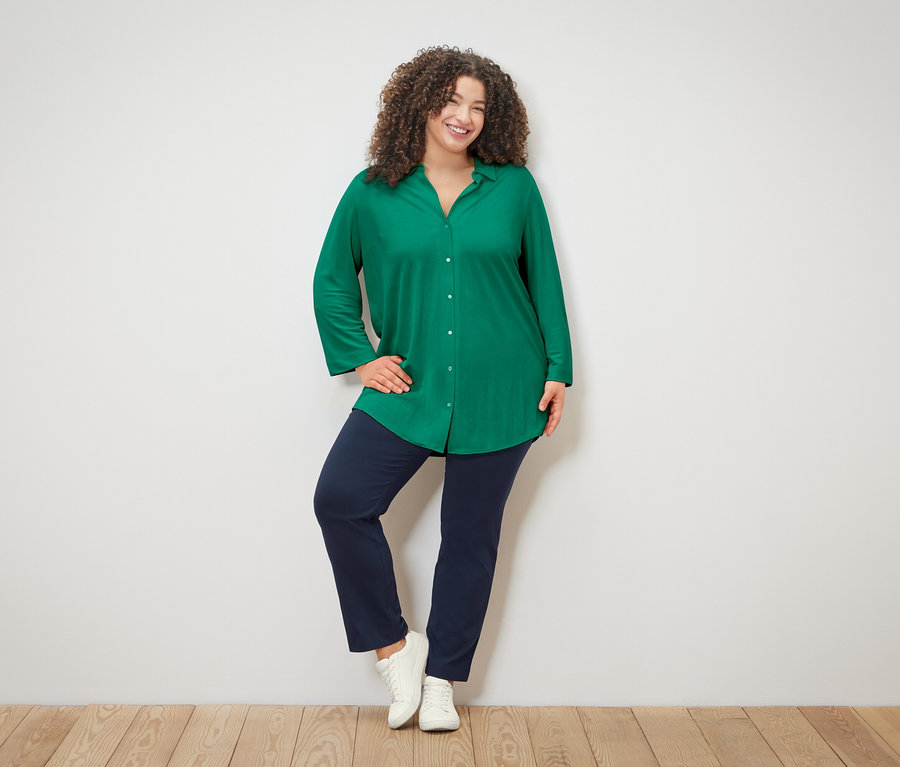 Une femme aux cheveux bouclés, portant un chemisier vert, un pantalon bleu et des baskets blanches, pose devant un mur blanc.