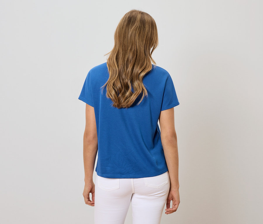 Vue de dos d'une femme aux cheveux blonds ondulés portant un t-shirt bleu et un jean blanc.