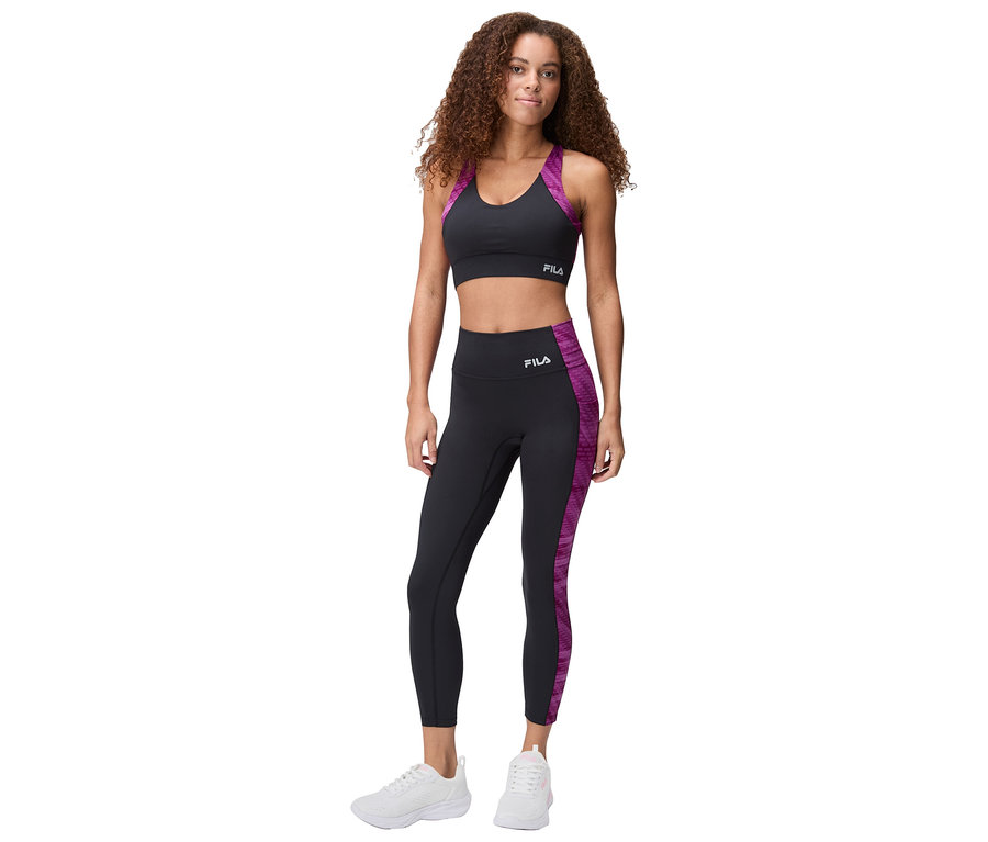 Femme debout en soutien-gorge de sport noir Fila avec des accents violets et un legging avec des baskets blanches.