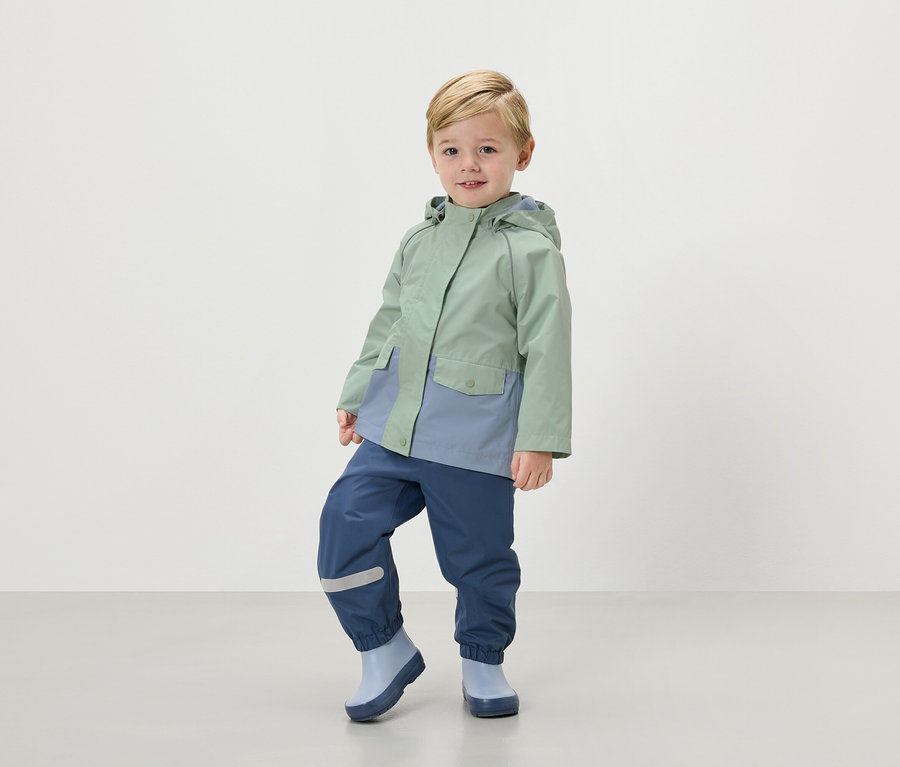 Un garçon blond porte un manteau de pluie vert menthe, un pantalon de pluie bleu marine et des bottes en caoutchouc bleu clair.