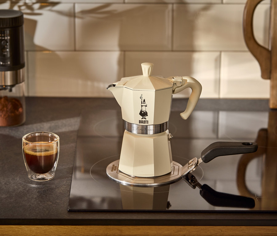 Une cafetière Bialetti Moka Express, 190 ml, crema est posée sur une plaque d’adaptation à induction Bialetti sur une plaque de cuisson. À l'arrière-plan, il y a une tasse de café.