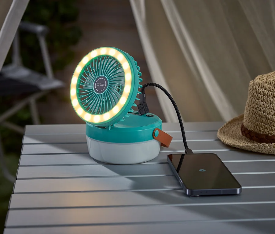 Un ventilateur de camping turquoise avec un anneau lumineux charge un smartphone sur une table de camping, à côté d'un chapeau de paille.