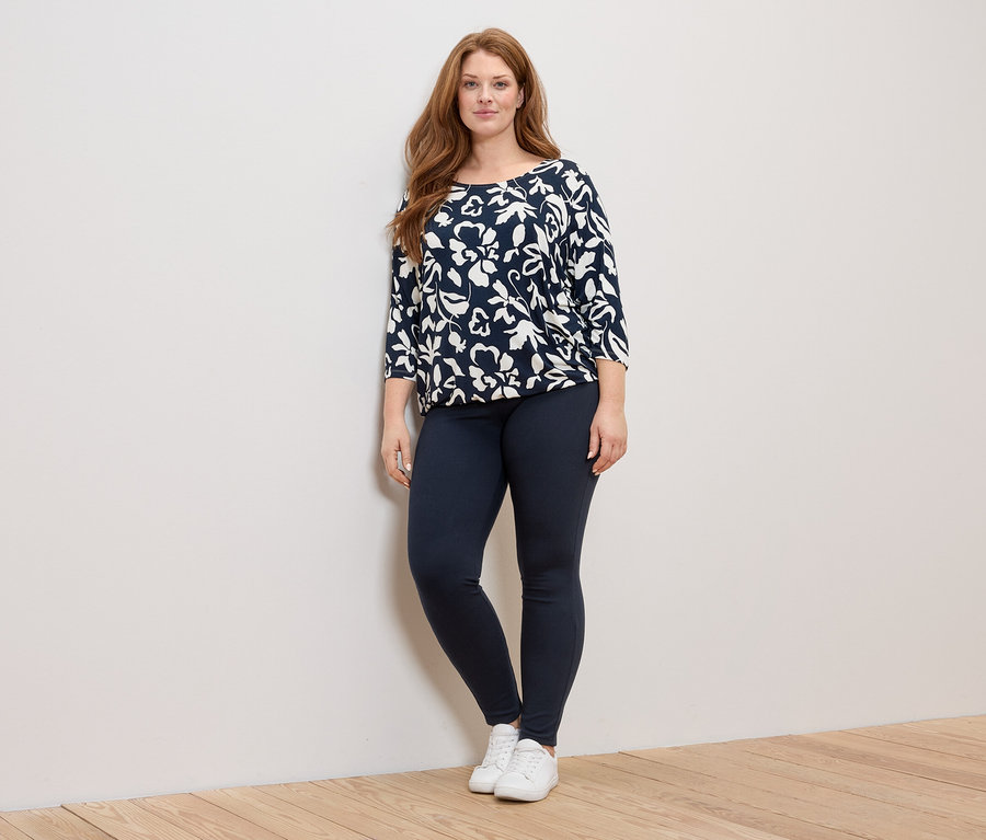 Une femme aux longs cheveux roux se tient debout, portant un haut bleu marine et blanc à motifs floraux, un legging bleu marine et des baskets blanches.