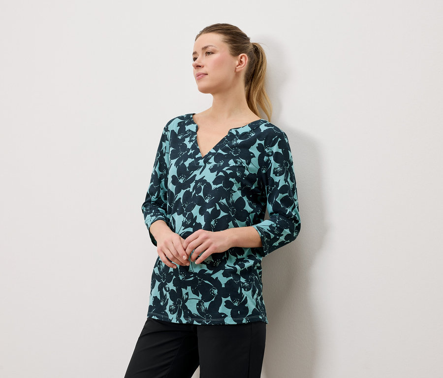 Femme aux cheveux blonds portant un chemisier bleu-vert à imprimé floral et un pantalon noir.