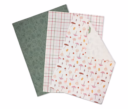Trois serviettes en tissu superposées : une vert olive à motifs de feuilles, une à carreaux et une blanche à motifs de champignons.