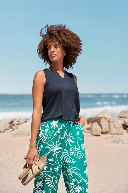 Une femme à la plage portant des Créoles Pavé et des Claquettes. Elle porte un haut foncé et un pantalon vert à motifs.