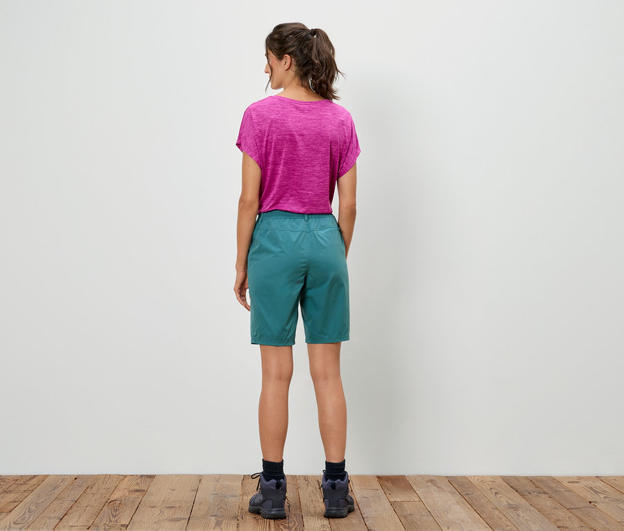 Une femme se tient dos à la caméra, elle porte un t-shirt rose et un short vert.