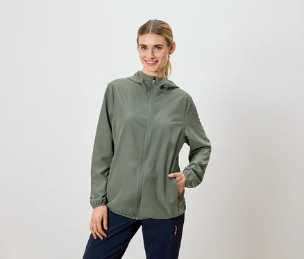 Une femme blonde porte une veste à capuche vert olive et un pantalon bleu marine.
