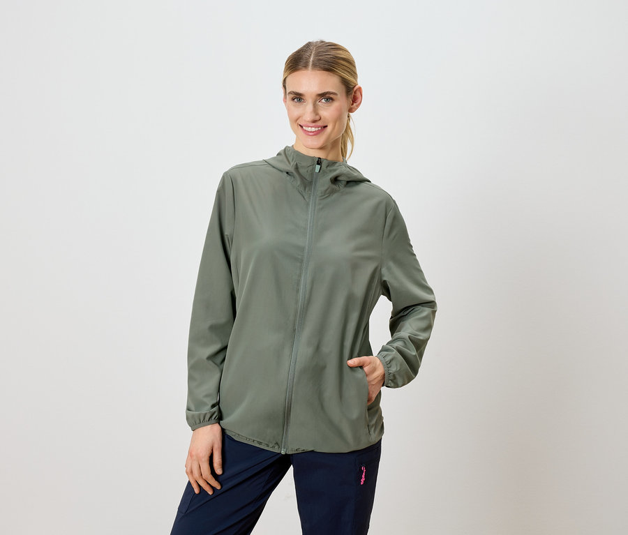 Une femme blonde porte une veste à capuche vert olive et un pantalon bleu marine.