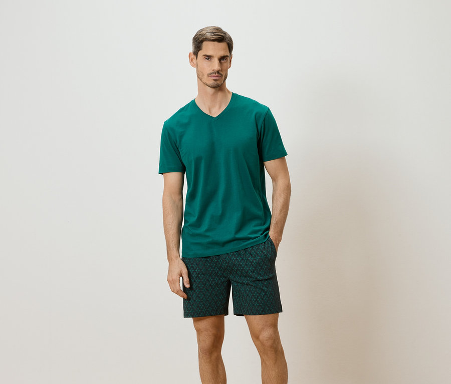 Homme portant un t-shirt vert émeraude et un short à motifs sur fond neutre.