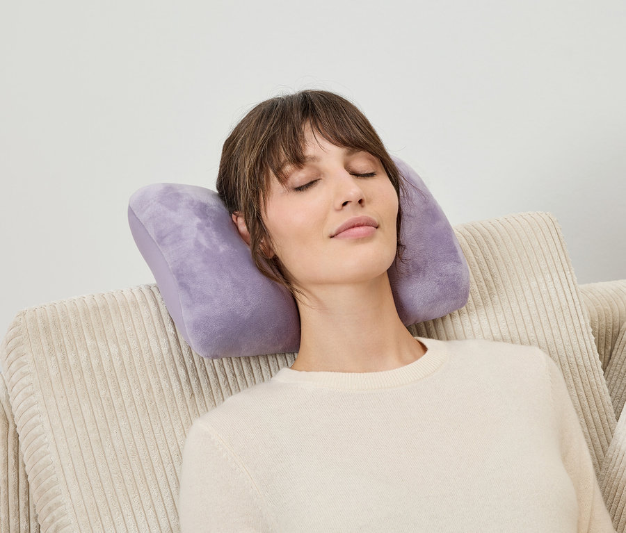 Une femme se repose les yeux fermés sur un canapé beige en velours côtelé et un coussin de nuque violet.