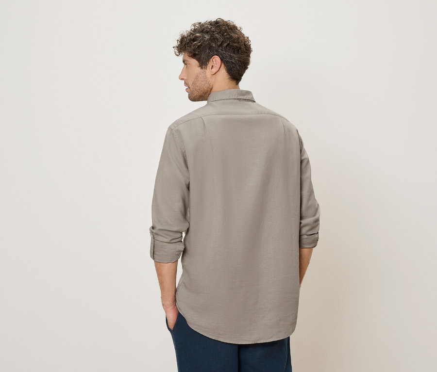 Un homme aux cheveux bouclés se tient dos à la caméra, portant une chemise grise et un pantalon bleu.