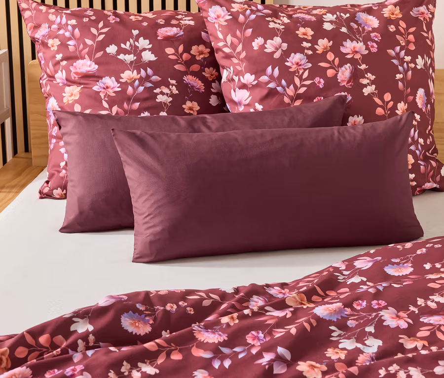 Sur un lit, deux oreillers bordeaux et deux taies d'oreiller à motifs floraux. Une couette bordeaux est posée au pied du lit.
