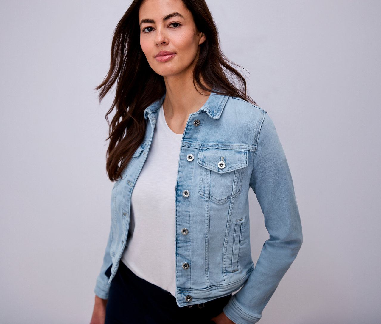 Une femme aux cheveux bruns portant une veste en jean bleu clair sur un t-shirt blanc.