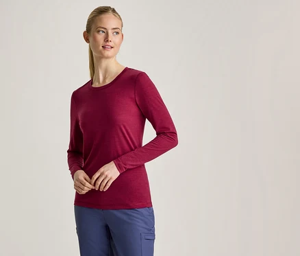 Une femme aux cheveux blonds attachés en queue de cheval porte un t-shirt bordeaux à manches longues et un pantalon bleu, debout les mains jointes devant elle.