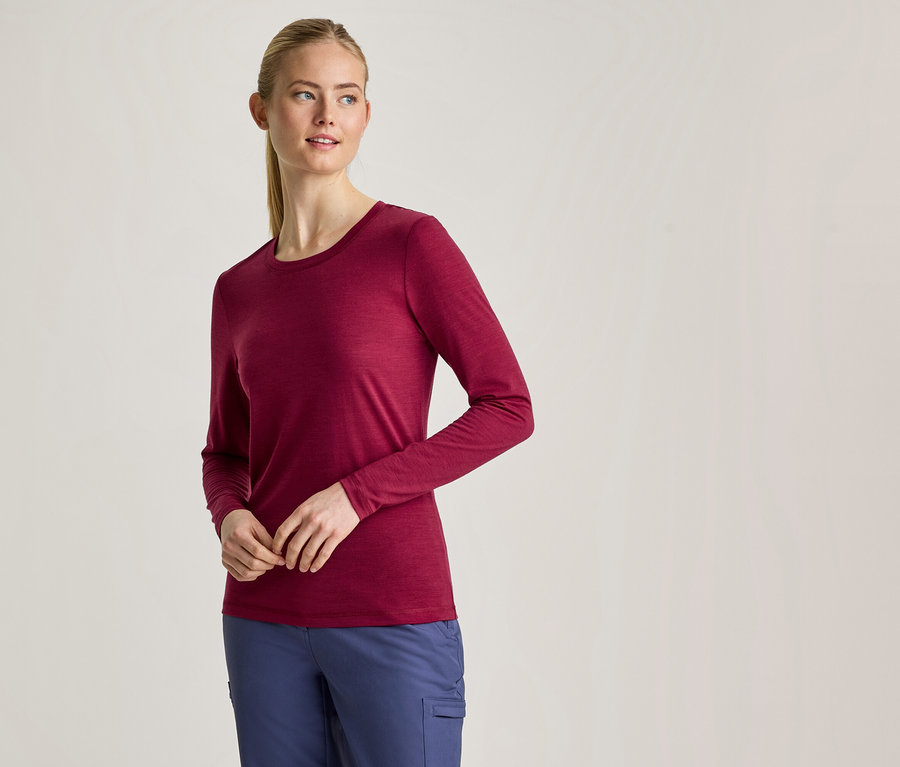 Une femme aux cheveux blonds attachés en queue de cheval porte un t-shirt bordeaux à manches longues et un pantalon bleu, debout les mains jointes devant elle.