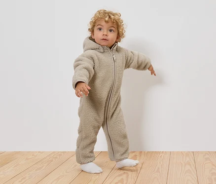 Un jeune enfant aux cheveux blonds bouclés porte une combinaison en teddy beige à capuche et des chaussettes blanches, debout sur un plancher en bois.
