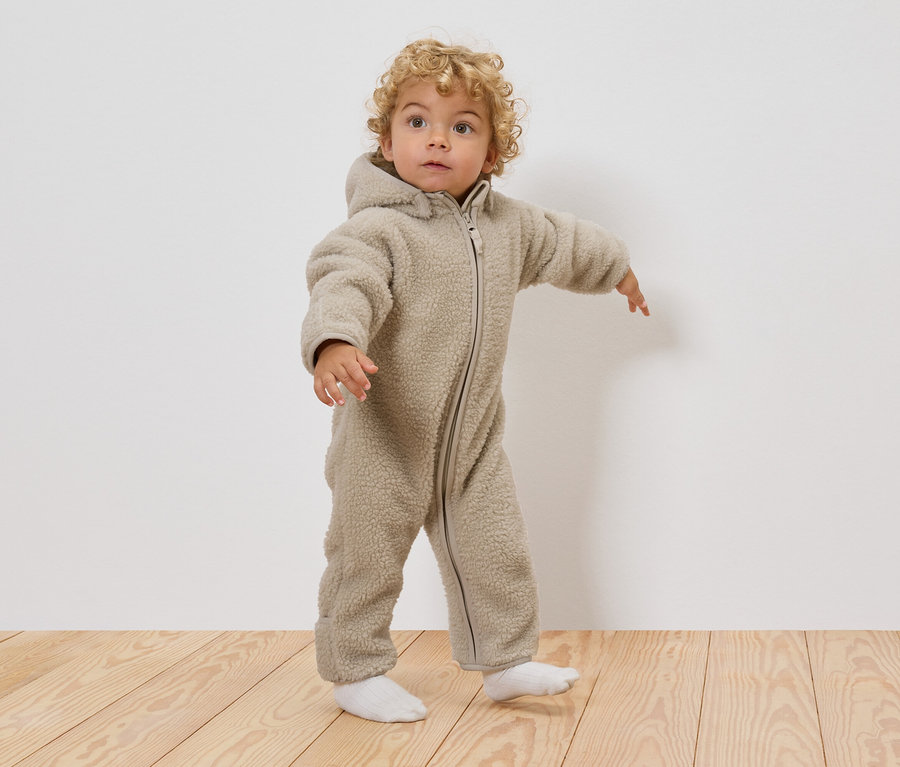 Un jeune enfant aux cheveux blonds bouclés porte une combinaison en teddy beige à capuche et des chaussettes blanches, debout sur un plancher en bois.
