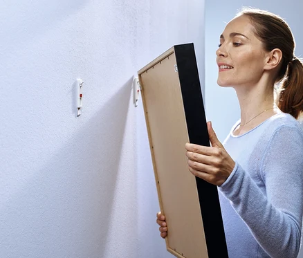 Une femme tient un tableau encadré devant un mur avec deux crochets adhésifs.