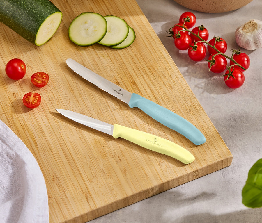 Sur une planche à découper en bois reposent une courgette entière, des tranches de courgette, des tomates cerises, une gousse d'ail et deux couteaux avec des manches bleu clair et jaune.