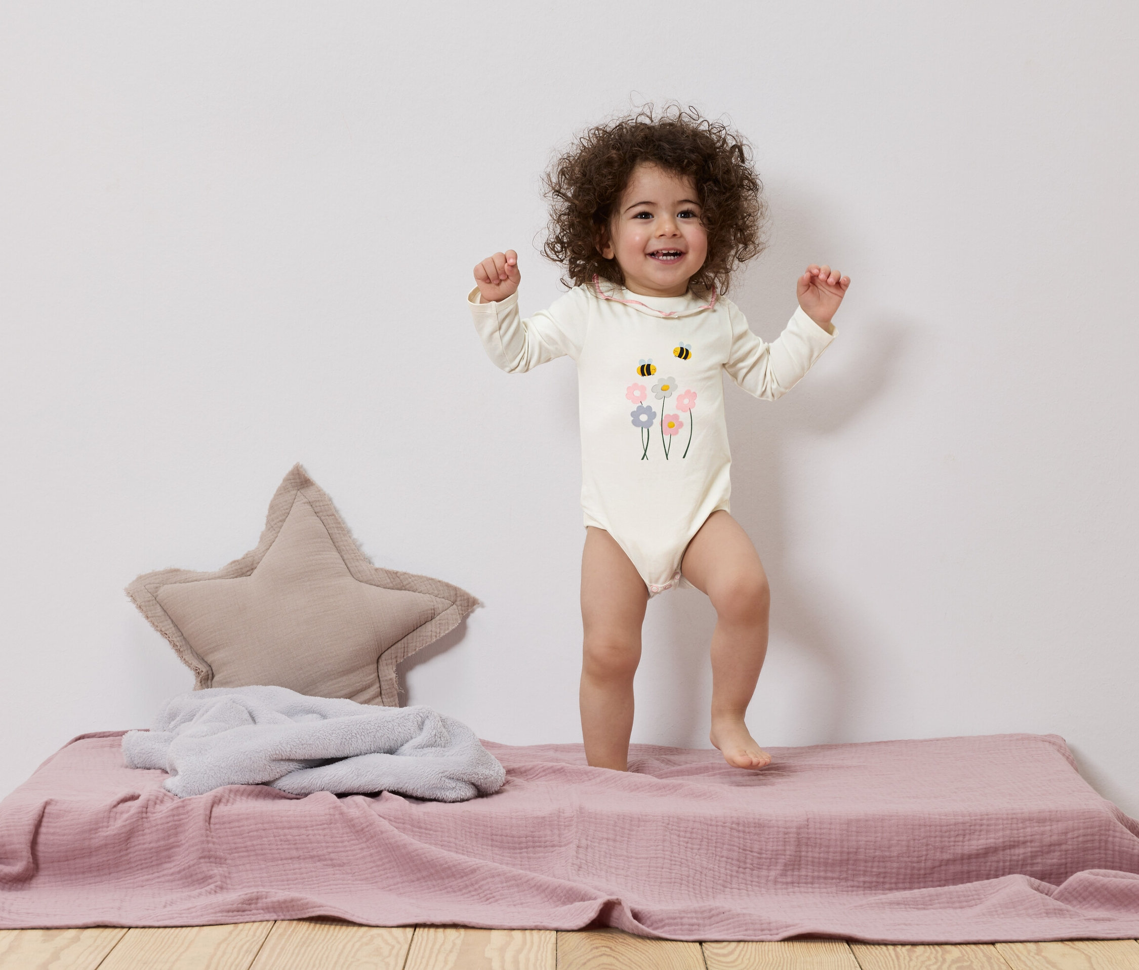 Un bébé souriant aux cheveux bouclés se tient debout sur une couverture de jeu rose à côté d'un coussin en forme d'étoile.