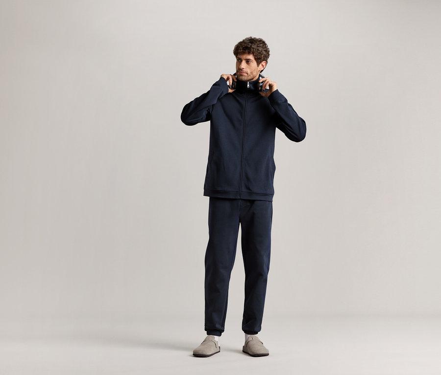 Un homme debout tenant des écouteurs à la main, portant un survêtement d’intérieur bleu marine.