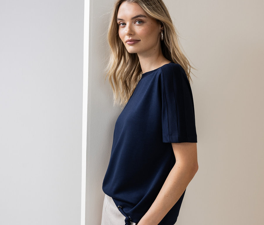 Femme appuyée contre un mur portant un t-shirt bleu marine et un pantalon clair.