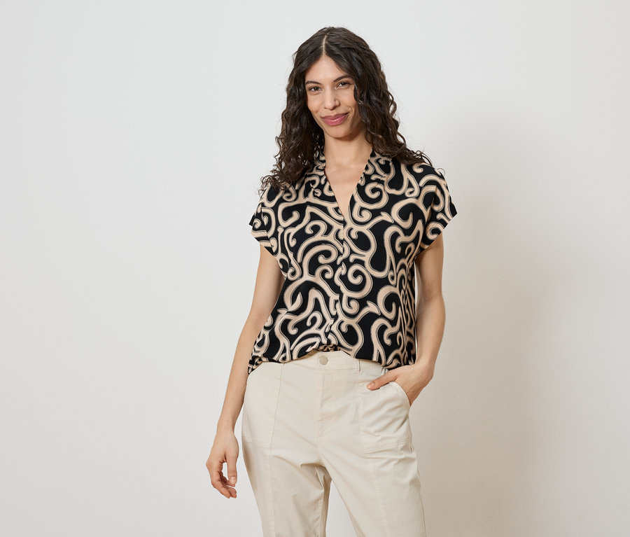 Une femme aux cheveux bouclés porte un chemisier noir à motifs beiges et un pantalon beige.