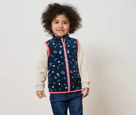 Un enfant souriant aux cheveux bouclés porte un gilet bleu marine à fleurs avec une fermeture éclair rose sur un haut beige et un jean.