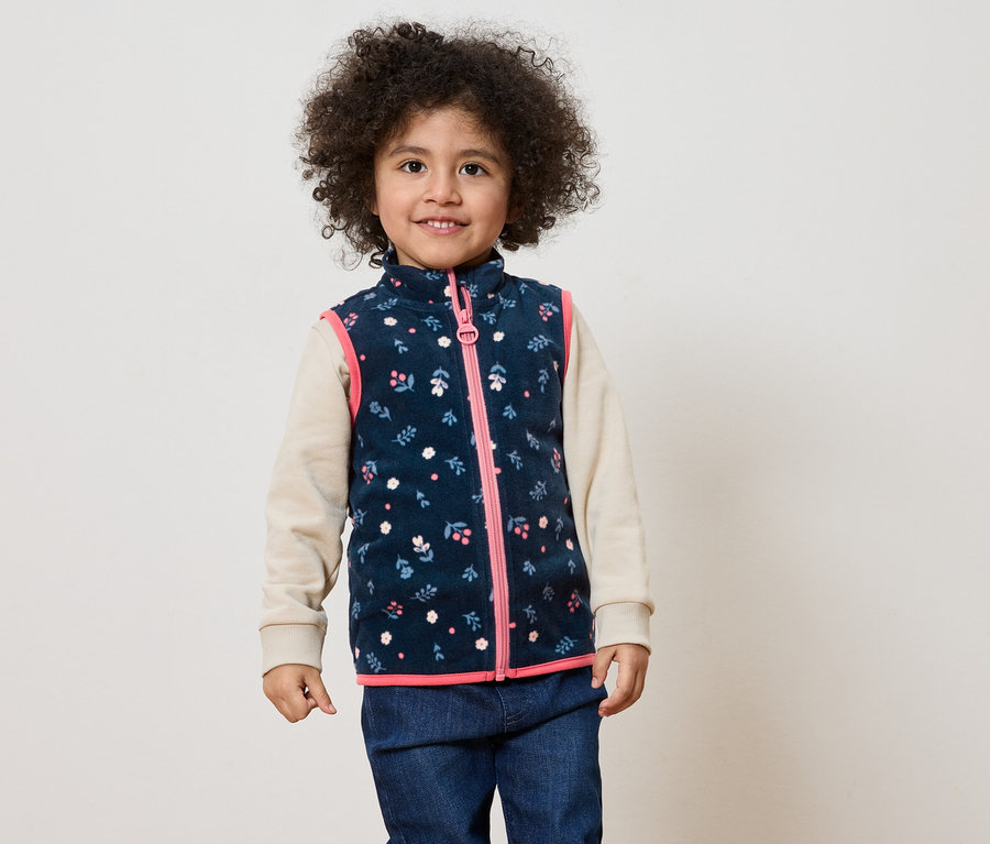 Un enfant souriant aux cheveux bouclés porte un gilet bleu marine à fleurs avec une fermeture éclair rose sur un haut beige et un jean.