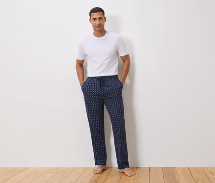 Un homme se tient devant un mur blanc, portant un t-shirt blanc et un pantalon de pyjama bleu marine à pois.
