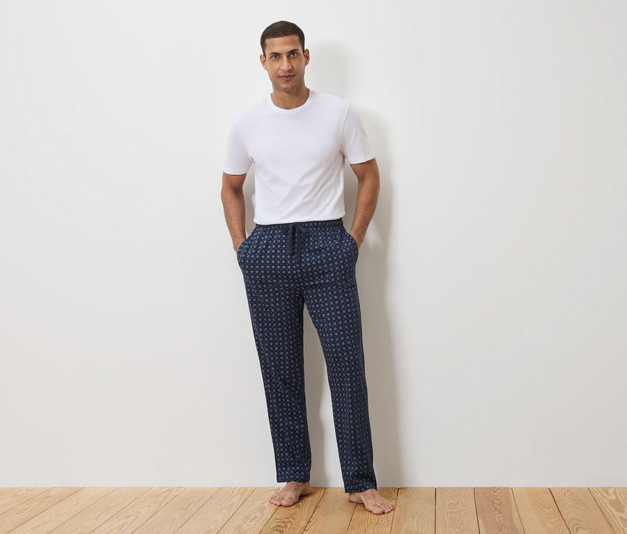 Un homme se tient devant un mur blanc, portant un t-shirt blanc et un pantalon de pyjama bleu marine à pois.