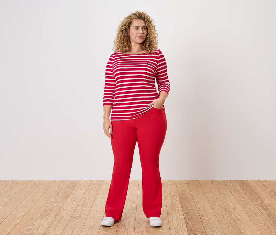 Femme debout, la main dans la poche de son pantalon, portant un haut rayé rouge et blanc, un pantalon rouge et des chaussures blanches.