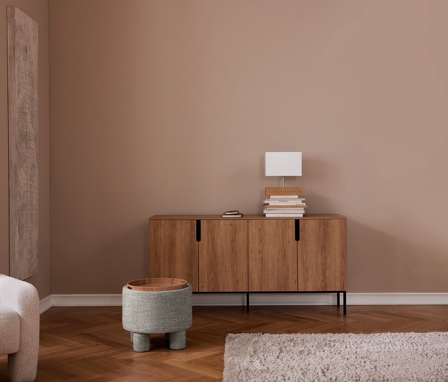 Un salon avec des murs marron clair, un parquet en chevrons et un tapis shaggy clair. Un buffet en bois avec une lampe rectangulaire blanche et des livres est adossé au mur. À sa gauche, un pouf rond gris avec un plateau en bois, et un fauteuil clair est partiellement visible à l'extrême gauche.